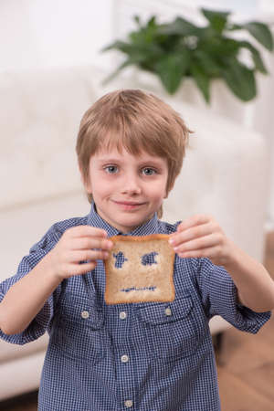 Portrait view of child boy sitting at home. blond boy showing bread at breakfastの写真素材