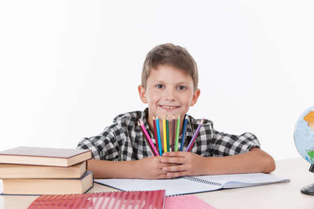 cute boy sitting at table and holding colorful pencils. handsome little schoolboy studying on white backgroundの写真素材