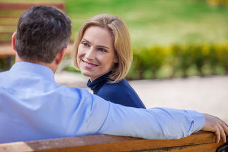 Adult smiling couple looking on each other sitting on bench. beautiful elegant mid age couple daydreaming outdoorsの写真素材