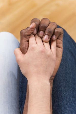 Close up of couple holding hands while sitting. top view of woman putting her hand on african man handの写真素材