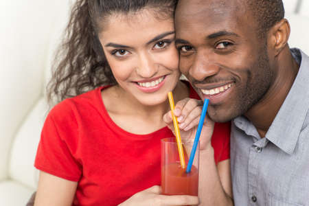 Young couple sharing glass of orange juice and looking at camera. black man and indian woman drinking juice with strawの写真素材