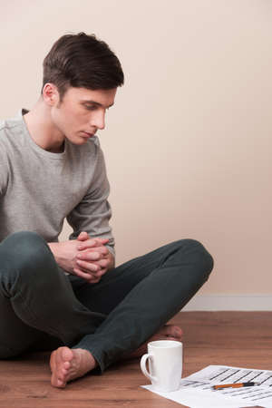 young man sitting on floor with legs crossed. handsome guy reading documents and drinking on wooden floorの写真素材