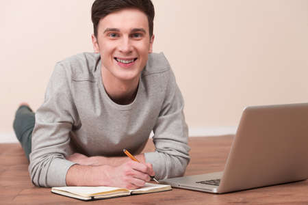 Cheerful young man lying on floor with notebook. Casual young man laying on floor at home, browsing internet, smiling.の写真素材
