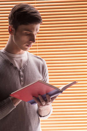 young male student reading book. closeup of man standing near window with jalousieの写真素材