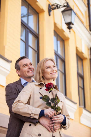 closeup shot of woman smiling and holding rose looking up. adult man standing behind woman and hugging herの写真素材