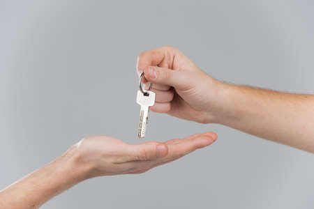 Male hand holding key and handing it over to another person. Handing key from one hand to another on grey backgroundの写真素材