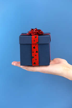 blue gift box in man hand on blue background. male holding box with red ribbon with hearts