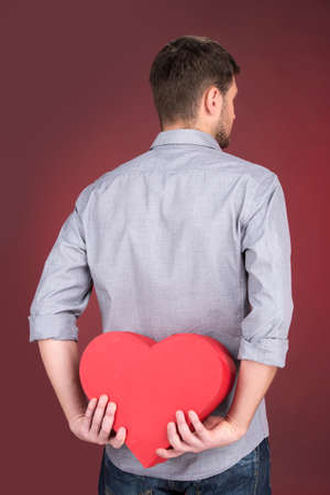 Portrait of young man holding heart shape over red background. back view of man hiding his valentine giftの写真素材