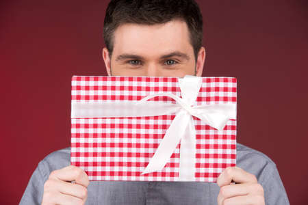 Present gift in hands of smiling man covering half face. closeup on happy man standing on red backgroundの写真素材