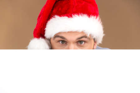 young man looking into camera with blank panel isolated on brown background. Smiling male hiding behind blank card and wearing red hatの写真素材