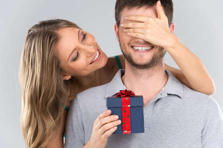 romantic woman covering her boyfriend's eyes. girl standing behind man with gift isolated on grey background