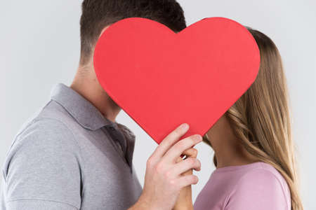 Two young dates behind heart with their faces close to one another. Happy couple kissing behind red heartの写真素材