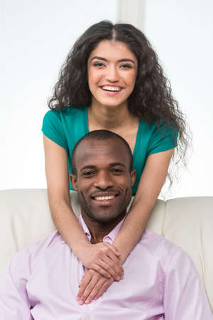 Indian girl standing over man sitting on sofa and hugging. black african man sitting on sofa and smilingの写真素材