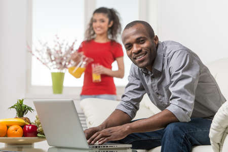 Handsome young African man sitting on couch and using laptop. girlfriend standing at background and drinking juiceの写真素材