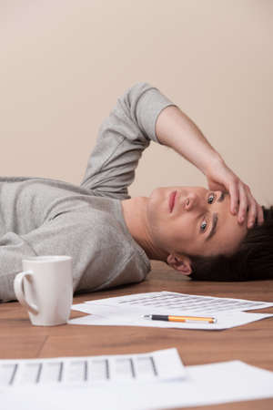 young man lying on floor and looking into camera. tired guy resting with documents and cup on wooden floorの写真素材
