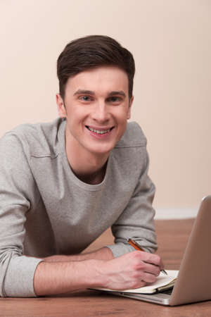 Cheerful young man lying on floor with laptop. Casual young man laying on floor at home, browsing internet, smiling.の写真素材