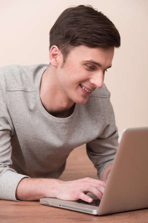 Cheerful young man lying on floor with laptop. Casual young man laying on floor at home, browsing internet and smiling.の写真素材