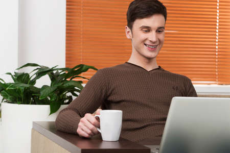 young man using laptop and drinking tea. Portrait of man working on laptop sitting at tableの写真素材
