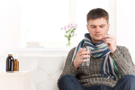 Sick caucasian man holding pills and water. Sick young man with flu drinks water on sofaの写真素材