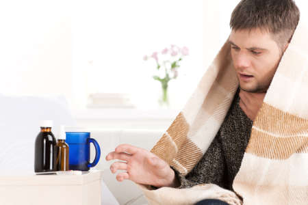 young man sitting on sofa covered with blanket. sick man looking tired and ill taking bottlesの写真素材