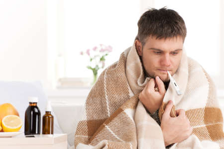Man with cold sitting on sofa with thermometer in mouth. Man at home sick with flu, taking his temmperatureの写真素材