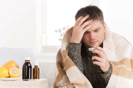Portrait of sick middle aged man with thermometer. Guy wrapped in plaid sitting on sofa is illの写真素材