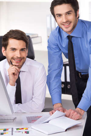 two businessmen having discussion in office. Two businessmen looking at camera and graphs on tableの写真素材