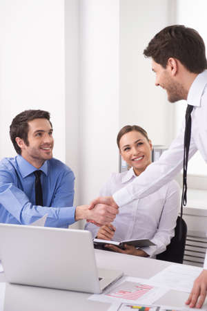 Side view of three people working on computer at office. Businesspeople sitting at desk in office and shaking handsの写真素材