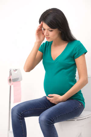 Young pregnant woman sitting in toilet. girl holding head and belly feeling headacheの写真素材