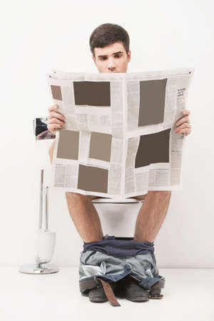 Young handsome man sitting on toilet and reading magazine. guy reading articles and stories in newspaper while in looの写真素材