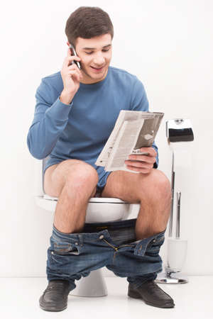 Young handsome man sitting on toilet and reading magazine. guy reading articles and talking on cell phoneの写真素材