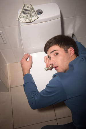 young man lying on toilet seat. drug addict kneeling over toilet seat with dollarsの写真素材