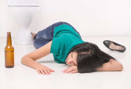 Drunk woman lying on toilet floor. black-haired girl sleeping on toilet tile with bottleの写真素材