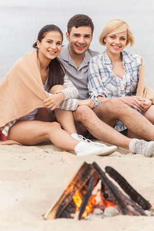 three friends sitting under blanket and warming near fireの写真素材