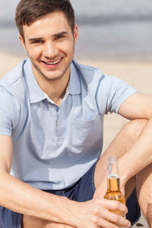 young man holding bottle of beer. smiling guy sitting on beach and looking into cameraの写真素材
