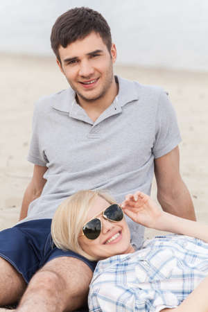 Attractive young couple sitting on beach at summer. happy woman lying on man's legs in glassesの写真素材
