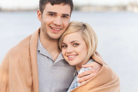 Attractive young couple standing on beach at summer. happy woman and man standing under blanket and smilingの写真素材
