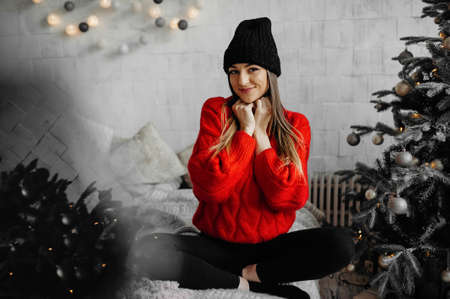 Photo of charming happy young woman in sweater with a black hat with standing cross-legged on a bed enjoying christmasの写真素材