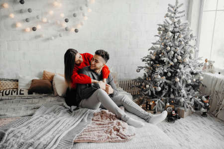 Happy couple in winter sweaters having fun lying on cozy bed and laughing, woman hugging her boyfriend spending winter holidays at home in warm christmas atmosphereの写真素材