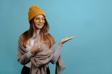 Portrait of stylish, pretty, smiling woman with scarf and winter yellow hat shows with her hands to the side at copy space, isolated on a turquoise background. Advertising conceptの写真素材