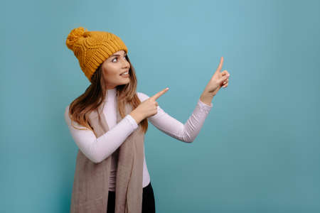 Portrait of stylish, pretty, smiling woman with scarf and winter yellow hat points his fingers up at copy space isolated on a turquoise background. Advertising conceptの写真素材