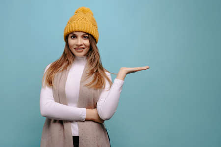 Portrait smiling woman with scarf and winter yellow hat shows with her hands to the side at copy space, isolated on a turquoise background. Advertising conceptの写真素材