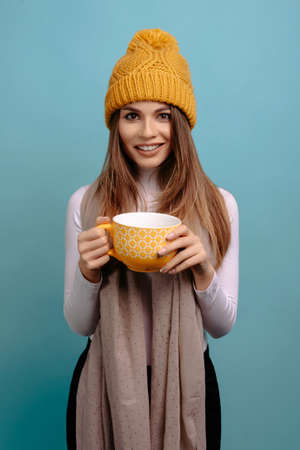 Woman holding yellow cup of tea wearing scarf and winter yellow hat, smiling at the camera over turquoise backgroundの写真素材