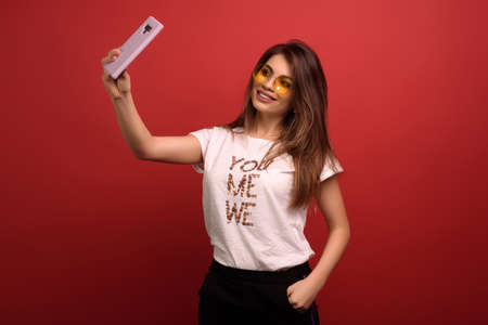 Portrait of lovely attractive happy woman making selfie isolated over red background, wearing white T-shirt and yellow glasses. Technology conceptの写真素材