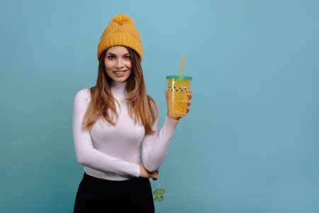 Portrait of stylish, pretty, smiling woman waring winter yellow hat, holds a yellow drinking glass in his hand, isolated on a turquoise background. Advertising conceptの写真素材