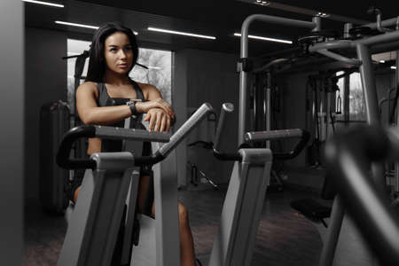 Fitness sport woman in the gym. Young sports girl during a break after working out. Caucasian woman sitting on the gym machine and looking at herself in the mirror, resting after training.の写真素材