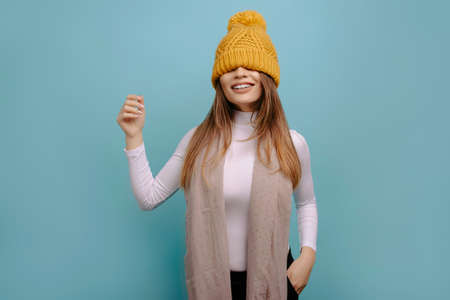 Portrait of playful, pretty woman having fun with the hat over his eyes smiling at the camera with his hand up, isolated on a turquoise background.の写真素材