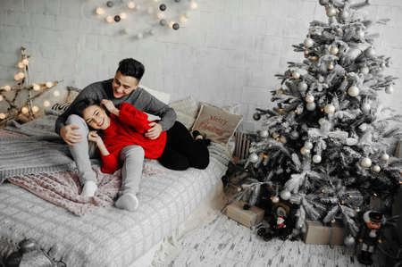 Cheerful couple in winter sweaters having fun lying on cozy bed and laughing , woman hugging her boyfriend spending winter holidays at home in warm and romantic atmosphere.の写真素材