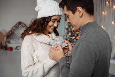 Charming young curly woman in winter hat excited with getting present from boyfriend. Happy couple with Christmas presents enjoying family cozy momentsの写真素材