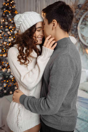 Lovely young couple hugging standing near decorated christmas tree celebrating Christmas at home. Woman and man enjoying perfect relationships and spending winter vacations in cozy home interiorの写真素材